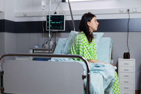 Hospitalized sick woman with illness recovering after medical surgery sitting on bed in hospital ward during recovery waiting for consultation. Patient healthcare treatment in modern clinicの写真素材