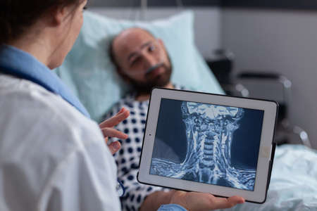 Specialist physician woman examining bones radiography using tablet computer discussing sickness symptom in hospital ward. Sick man patient with nasal oxygen tube resting in bedの写真素材