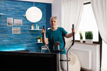 Focused senior woman doing workout training body muscle watching online video gym exercise on television using slimming bicycle at home in living room. Fitness pensioner working at cardio resistanceの写真素材