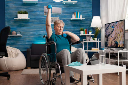 Disabled old woman in wheelchair raising arm training muscles resistance using dumbbells recovery after muscular disability watching cardio video on laptop. Pensioner doing gym exercise in living roomの写真素材
