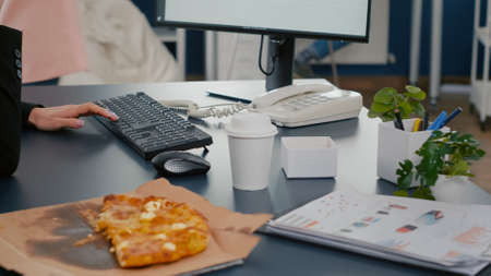 Closeup of businesswoman sitting at desk in front of computer eating pizza slice while talking at...