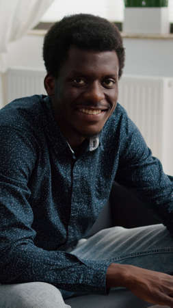 Portrait of black man in living room typing on laptop, looking up at the camera and smiling. African man working from home, computer user in flat apartment using social media freelancer in slow motionの写真素材