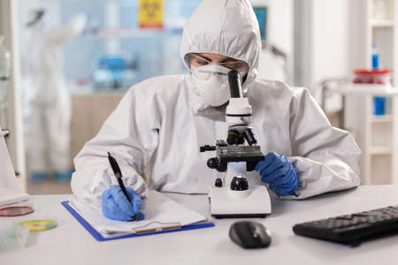 Medicine scientist dressed in ppe suit taking notes during vaccine research using microscope. Chemist in coverall working with various bacteria, tissue blood samples for antibiotics research.の写真素材