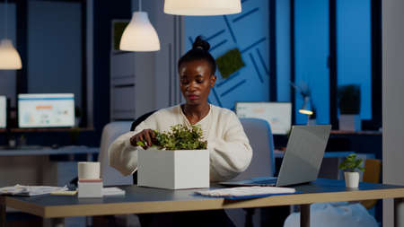 Sad african fired businesswoman packing things in box being dismissed from job late at night. Overworked unemployed woman closing laptop and leaving workplace office in midnightの写真素材