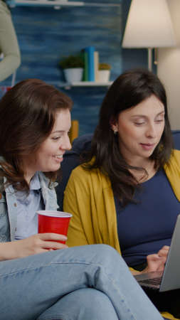 Multi-ethnic women enjoying time together watching at movie on laptop while resting on couch. In...