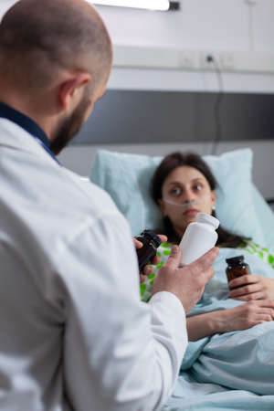 Sick woman resting in bed during medical examination wearing nasal oygen tube discussing with specialist doctor pain treatment in hospital ward. Physician man explaining pills medicationの写真素材
