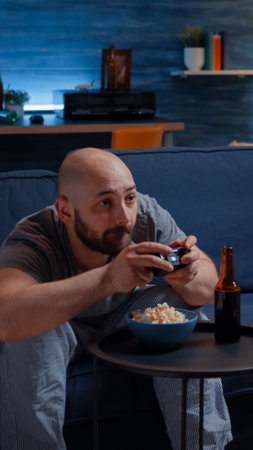 Competitive determined man playing videogame in living room at night focused eating popcorn enjoying free time. Involved gamer sitting on couch, playing video games using joystick, wireless controllerの写真素材