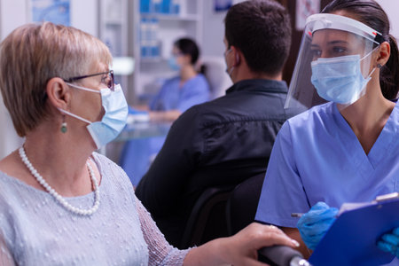 Close up of assistant explaining diagnosis to paralyzed old woman in wheelchair pointing on clipboard. Nurse wearing face mask against infection with coronavirus in hospital waiting area consultingの写真素材