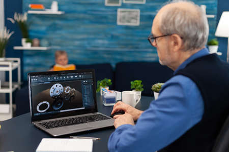Senior engineer working on deadline sitting at desk. Experienced engineer working from home office on mechanical project using laptop computer technology and wife is reading a book on sofa.の写真素材