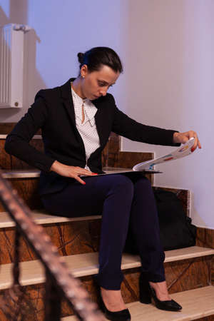 Exhausted tired overworked businesswoman holding financial reaport of deadline project. Serious entrepreneur working on job project sitting on staircase of business building at night hours for job.の写真素材