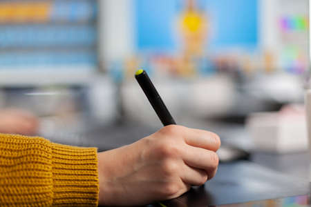 Close up on hand of young woman photo editor drawing on graphic tablet editing customer photo on computer with two monitors. Retoucher holding stylus pen retouching assets in creative agency officeの写真素材