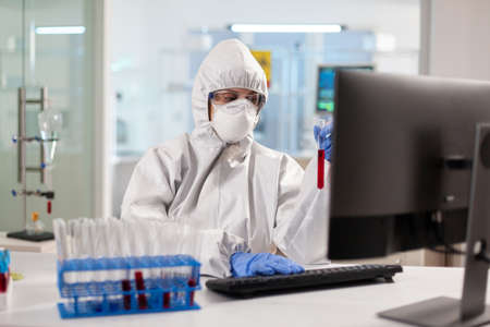 Medical researcher dressed in ppe suit looking at blood sample using computer. Scientist examining...