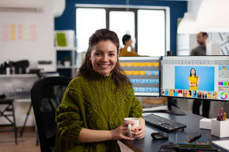 Woman retoucher looking at camera smiling sitting in creative media agency retouching client photos on PC with two displays. Photographer graphic editor working in start up office with digital assetsの写真素材