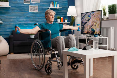 Handicapped senior woman in wheelchair stretching arms muscles exercising body resistance using workout dumbbells in living room. Handicapped pensioner looking at lifestyle aerobics video on tabletの写真素材
