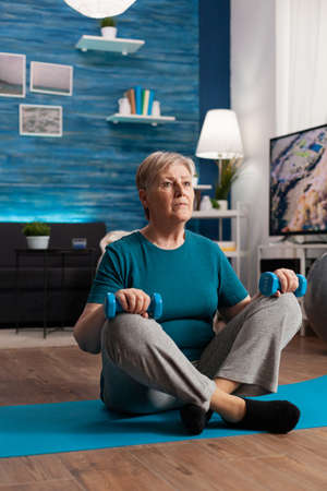 Senior woman in sportwear warming up abdominals muscles practicing gym body exercise using workout dumbbells. Pensioner sitting on yoga mat in lotus position during wellness routine in living roomの写真素材