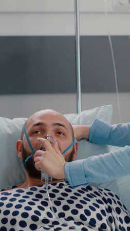 Close up of sick man patient resting in bed while doctor putting oxygen mask monitoring respiratory illness in hospital ward during healthcare emergency. Physician analyzing heartbeat pluseの写真素材