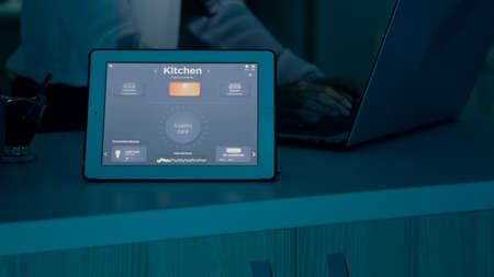 Female typing on laptop working from home with automation lighting system using voice controlled on tablet switching on bulbs. Smart gadget responds to commands, woman using automated app softwareの写真素材