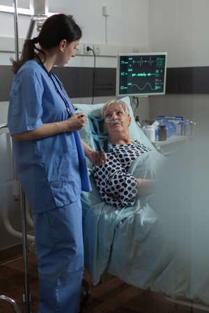 Elderly patient in radiology hospital room laying in bed listening nurse dicussing about chest x-ray radiography for respiratory diagnosis. Breathing with help from oxygen mask.の写真素材