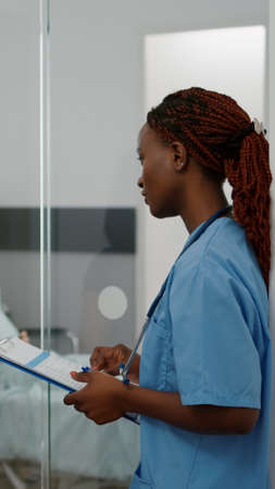 African american practitioner nurse monitoring sickness symptom explaining medical treatment to worried mother during clinical consultation. Hospitalized child resting in bed playing games on phoneの写真素材