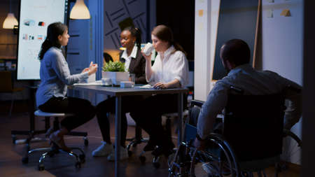 Focused overworked handicapped businessman in wheelchair sharing financial paperwork statistics overworking in business office meeting room. Diverse multi-ethnic brainstorming company ideas in eveningの写真素材