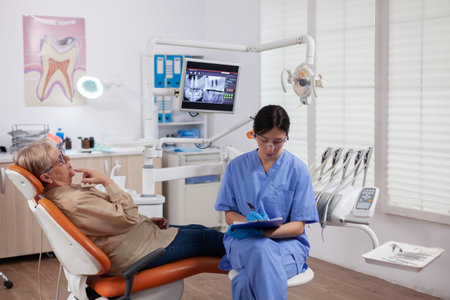 Assistant in dentist cabinet during elderly woman oral hygiene check up sitting on chair. Senior woman talking with medical nurse in stomatology office about teeth problem.の写真素材