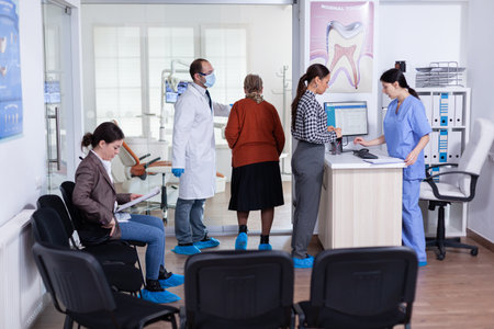 Young lady asking informations filling in stomatological form while patients talking sitting on chair in waiting area. People speaking in crowded professional orthodontist reception office.の写真素材