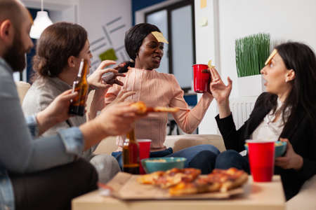 African american woman playing guessing game with friends after work at office. Multi ethnic group of workers guess imitation for fun cheerful activity while eating and drinking beerの写真素材