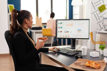 Businesswoman having delivery food order on desk during takeout lunchtime working in business company office. Entrepreneur woman eating takeaway lunch pizza slice management statistics.の写真素材
