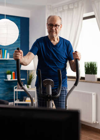 Active senior man training legs muscle resistance cycling bicycle machine in living room during lifestyle healthy workout. Pensioner watching fitness video on televison for well beingの写真素材