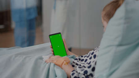Little kid holding mock up green screen chroma key phone with isolated display during sickness consultation in hospital ward. Sick child lying in bed recovering after disease infectionの写真素材