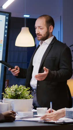 Focused man leader explaining management project using monitor working in company meeting office room late at night. Overworked diverse multiethnic teamwork brainstorming corporate strategy in eveningの写真素材