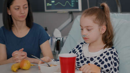 Mother standing with sick girl while eating healthy food breakfast during sickness expertise in hospital ward. Hospitalized girl having nutrition food recovering after medical surgeryの写真素材