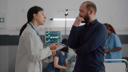 Pediatric woman doctor explaining disease symptoms to father while black nurse writing medical sickness treatment on clipboard in hospital ward. Sick little child resting in bed during examinationの写真素材