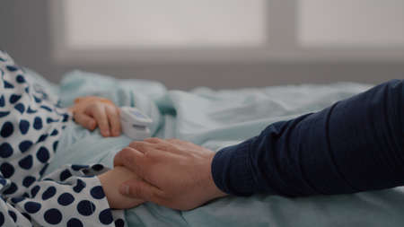Closeup of sick child resting in bed with oximeter on finger monitoring heartbeat pulse after suffering recovery surgery. Father holding daughter hands during sickness examination in hospital wardの写真素材