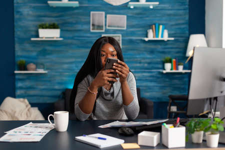 Black student looking into camera while socializing with friends group typing message on modern smartphone. African american woman sitting desk in living room browsing on social mediaの写真素材