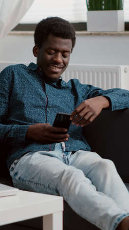 African american man with phone in hands looking at online streaming services content, enjoying working from home, connection via internet web services. Home technology userの写真素材
