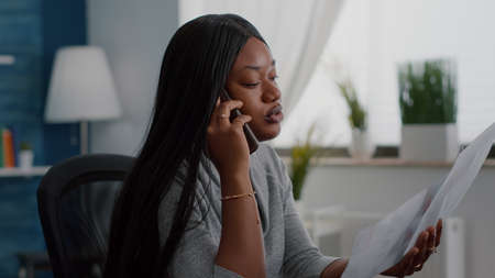 Upset black student sitting at desk explaining financial homework discussing with collegue using smartphone. Black woman working remote from home at online webinar using academic elearning platformの写真素材