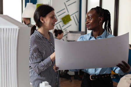 Colleagues architects women checking plans of blueprints layout design. Multi ethnic team of professional workers standing at building model maquette for modern technology projectの写真素材
