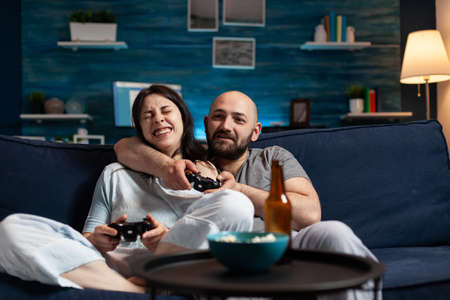 Determinated enthusiastic couple winning video games, hugged each other sitting on sofa playing, excited scream enjoying online soccer competition. Competitive happy gamers using gaming joysticksの写真素材