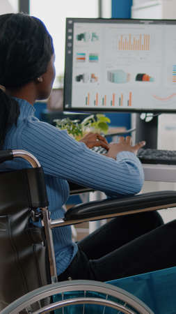 Busy focused african invalid paralized woman manager analysing graphics and financial statistics from computer, typing working in start-up business office sitting in wheelchair using modern technologyの写真素材