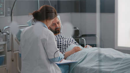 Professional doctors discussing sickness symptom with sick man writing medical tratment on clipboard working in hospital ward. Patient resting in bed while practitioner monitoring disease recoveryの写真素材