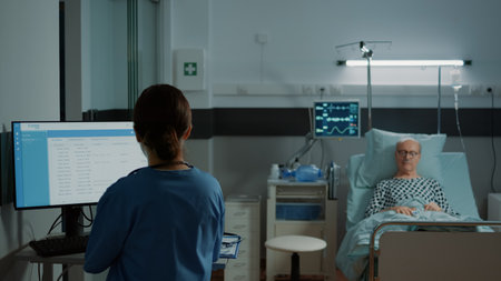 Nurse examining medical results on computer for patient laying in hospital ward bed. Sick old man waiting for intensive care information for healing disease, illness, emergencyの写真素材