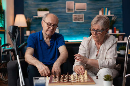 Senior man and woman focused on board game match relaxing at home. Old married couple playing chess together while enjoying retirement time on living room couch with crutches and walk frameの写真素材