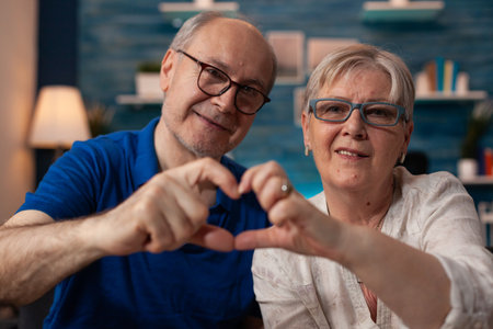 Portrait of retired man and woman holding hands in heart shape for romantic moment in living room. Happy senior couple showing love concept affection sign shape gesture sitting on couchの写真素材