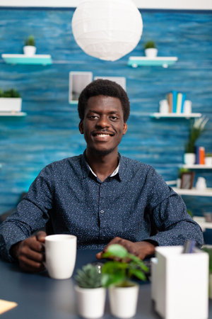 Portrait of african american man smiling into the camera at office desk. Black young man in modern flat apartment working from home using computer technology for digital businessの写真素材