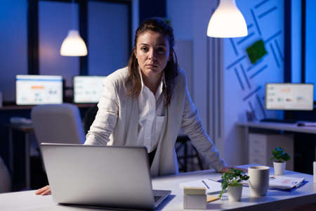 Business woman looking tired at camera standing near desk in start-up business company late at...