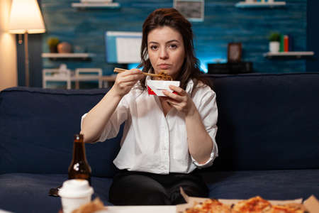 Portrait of caucasian female looking into camera while eating chinese chopstick relaxing on sofa in living room late at night. Woman enjoying takeaway food home delivered. Tasty fastfood lunch orderの写真素材