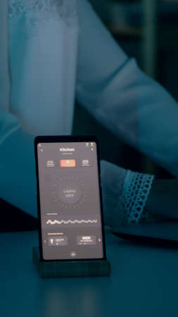 Woman typing at laptop sitting in home with automation lighting system using voice controlled on smartphone turning on bulbs. Speaker gadget responds to commands, person controlling ambiance lightの写真素材