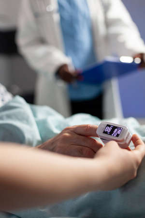 Close up of oxymeter attached on sick senior patient laying in hospital bed, monitoring hemoglobin. Medical staff using modern technology for reading blood oxygen saturation.の写真素材