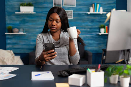 African american woman holding cup of coffee in hands socializing with friend using smartphone...